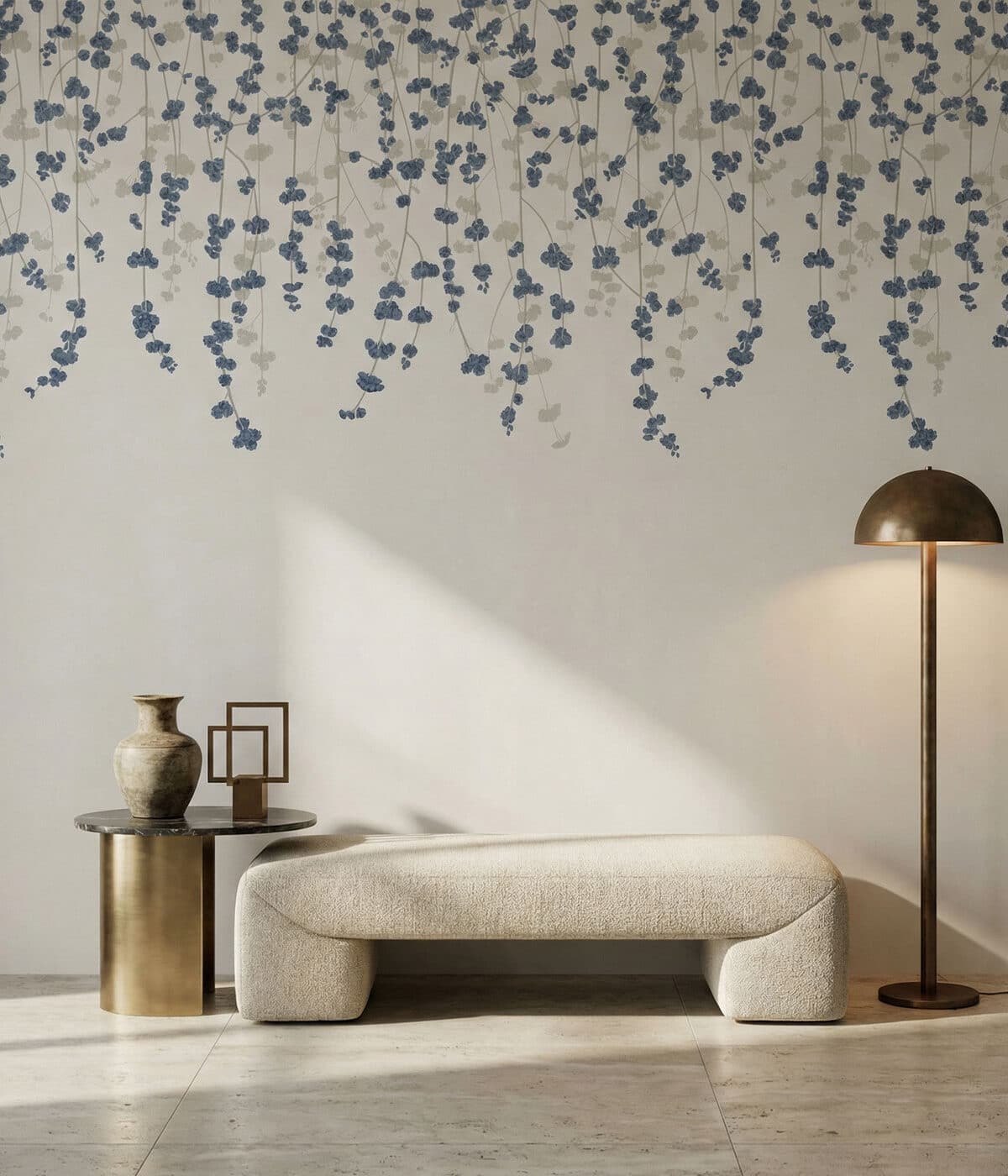 A minimalist interior with a soft white wall decorated along the top with cascading blue cherry blossom branches. Below, a low, curved cream boucle bench sits on a light stone floor. To the left of the bench is a small round side table with a brass base and dark top, holding a rustic ceramic vase and a geometric metal sculpture. To the right stands a slim brass floor lamp with a dome-shaped shade. Sunlight casts a diagonal beam of light across the wall, creating a calm and elegant atmosphere.