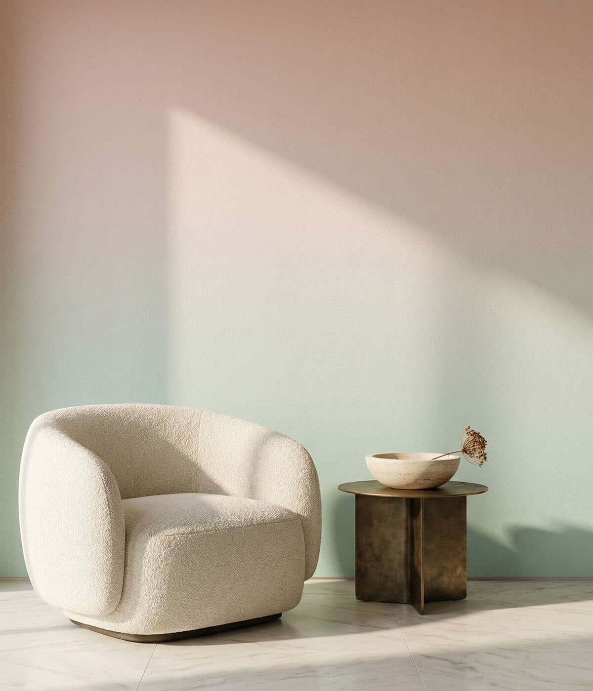A minimalist interior featuring a rounded cream boucle armchair beside a small brass side table holding a shallow stone bowl with a dried plant stem. The wall behind displays a soft ombre wallpaper that fades from warm pink at the top into pale teal at the bottom. Sunlight creates a large triangular shadow across the wall, enhancing the calm, airy atmosphere.