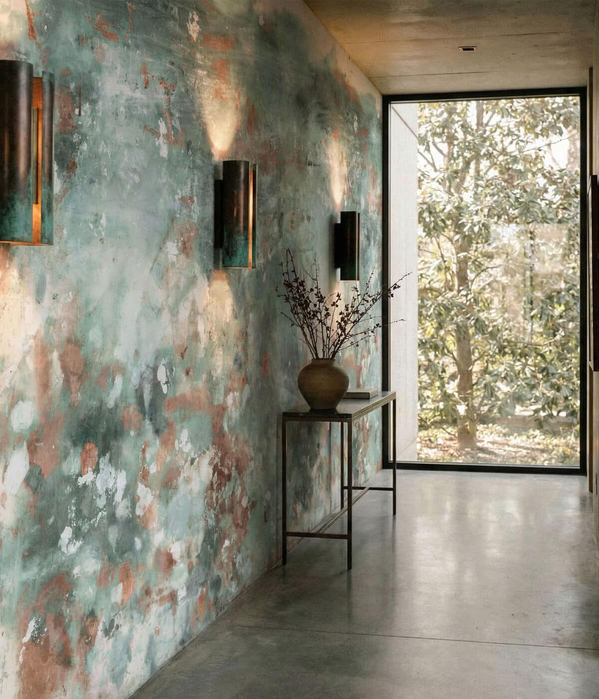 A modern hallway featuring distressed green and brown textured wallpaper with an abstract, weathered finish. Minimalist wall sconces cast warm light across the wall, while a slim console table with a ceramic vase sits beside a large window overlooking leafy trees outside.