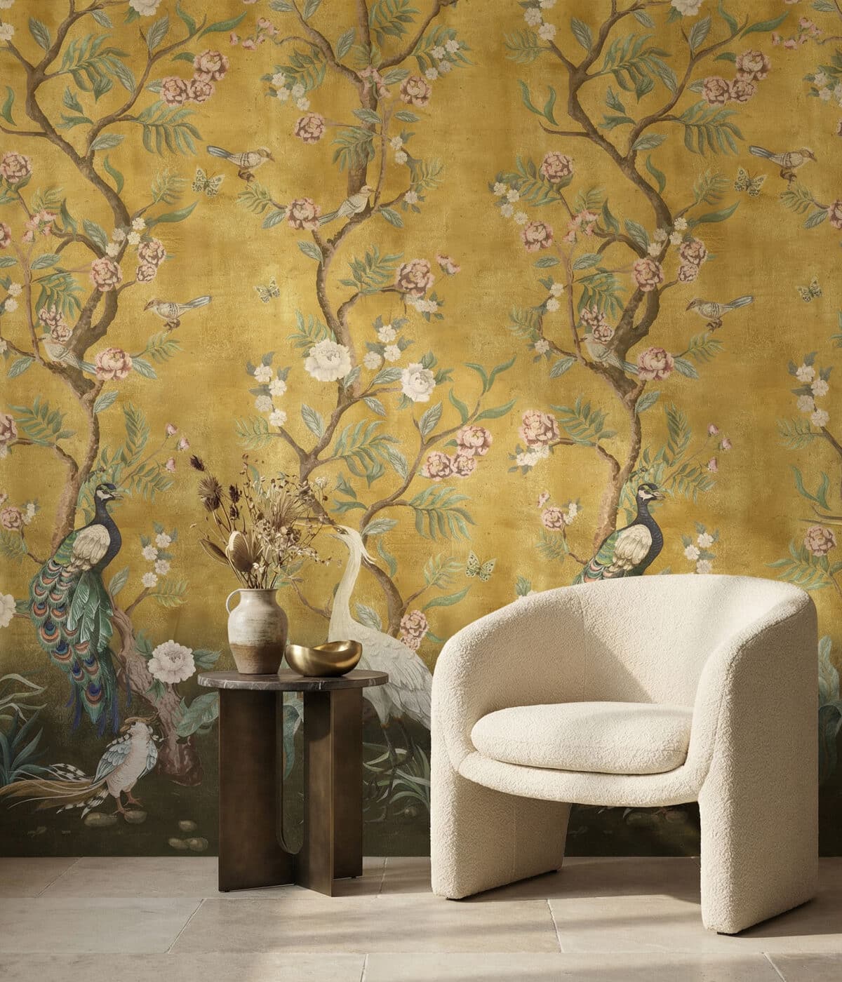 An elegant living space featuring golden chinoiserie wallpaper with delicate flowering branches, birds, and peacocks. A soft cream armchair sits beside a small bronze side table holding a vase with dried flowers, creating a warm and refined interior scene.
