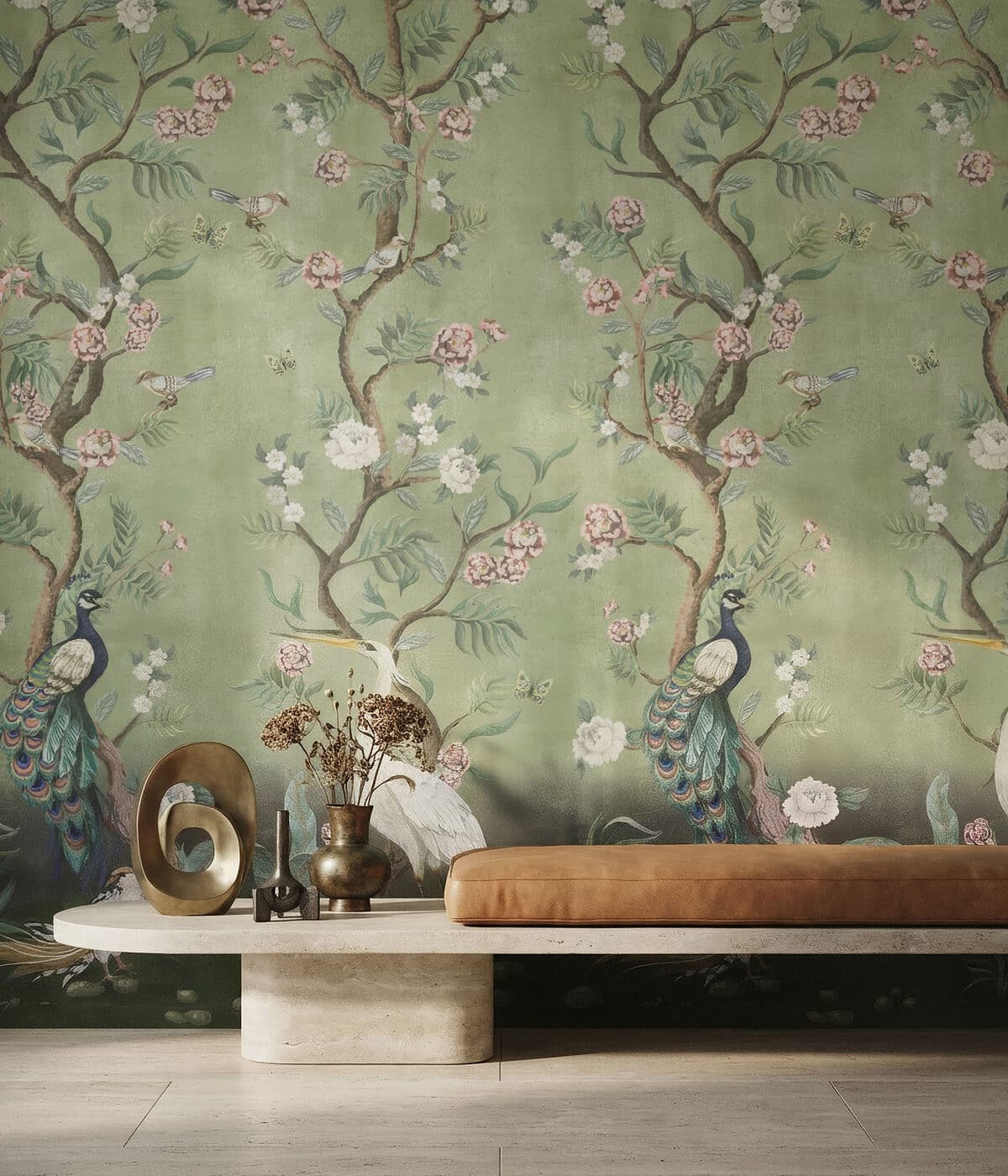 A stylish interior featuring a long stone bench topped with a caramel-brown leather cushion. Decorative objects—including a brass sculptural ornament and small metal vases with dried flowers—are arranged on the bench. The wall behind is covered in a green chinoiserie-style wallpaper depicting flowering tree branches, delicate blossoms, butterflies, and birds, including peacocks and a white crane, creating a lush, elegant garden scene.