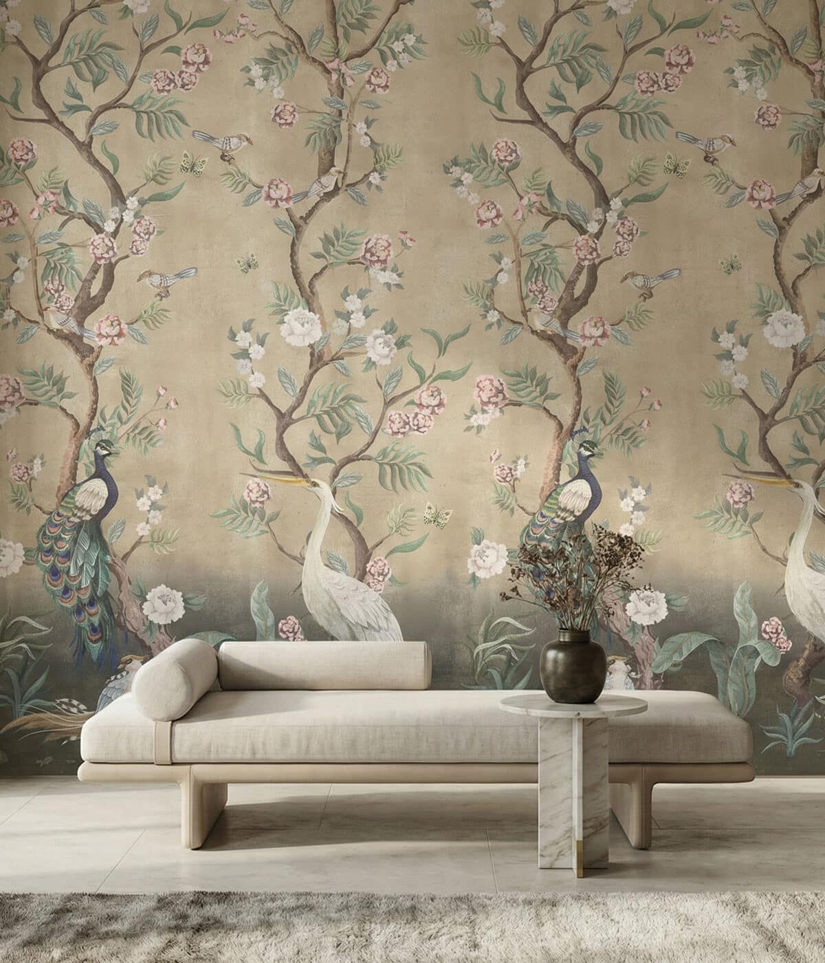 A contemporary living space featuring a light upholstered daybed with a cylindrical bolster cushion, placed in front of a cream-toned chinoiserie wallpaper. The wallpaper depicts elegant flowering tree branches with blossoms, butterflies, and birds including peacocks and a white crane. A small marble side table with a dark vase holding dried flowers sits beside the sofa, while the soft neutral furnishings contrast with the detailed decorative mural behind them.