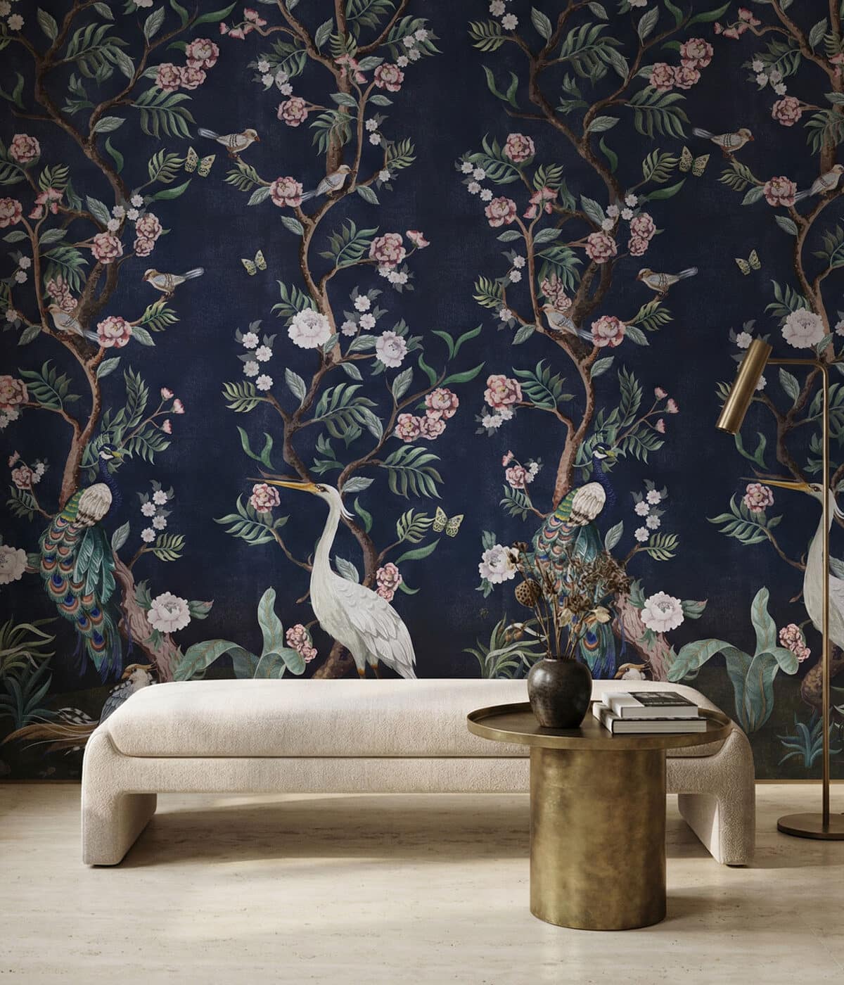 A refined interior with a cream upholstered bench set against a deep navy chinoiserie wallpaper featuring flowering tree branches, delicate blossoms, butterflies, and birds including a white crane and colorful peacocks. In front of the bench sits a round brass side table holding a small vase with dried flowers and a few books, while a slim brass floor lamp stands to the side. The dark wallpaper creates a rich, dramatic backdrop for the light furniture.