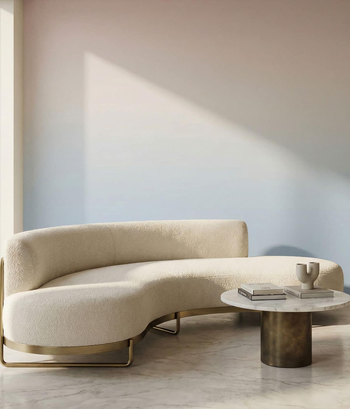A modern living room featuring a curved cream boucle sofa with brass frame accents and legs. In front of it sits a small round coffee table with a marble top and dark metal base, holding a couple of books and a sculptural ceramic vase. The wall behind the sofa is covered in a soft ombre wallpaper that fades from pale pink at the top into light blue at the bottom, while sunlight casts a large triangular shadow across the wall.