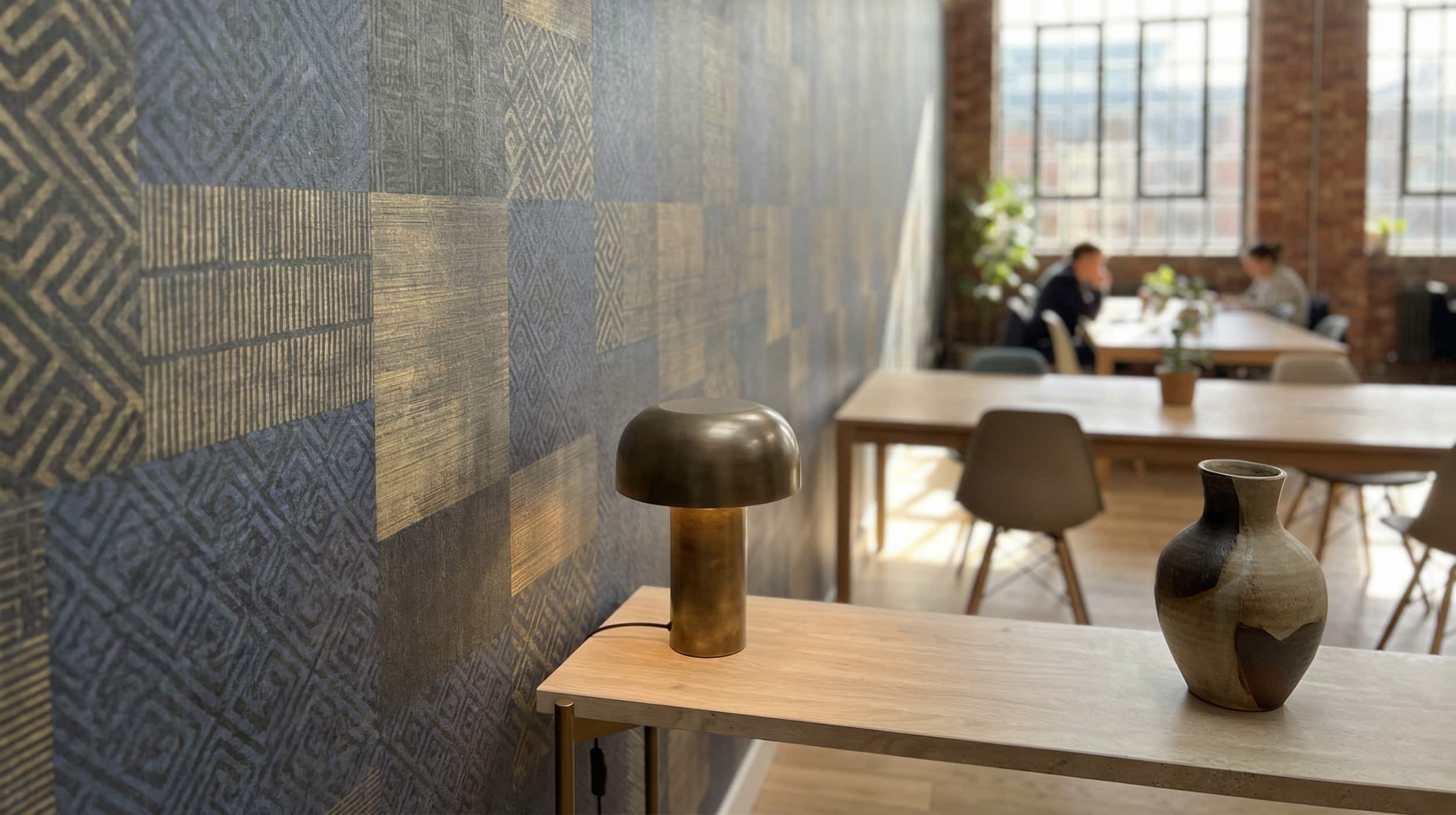 A modern, industrial-style office space features a large accent wall with a sophisticated geometric mural. The design consists of a grid-like pattern of large squares in navy blue and slate gray, overlaid with shimmering gold textures including horizontal stripes and intricate labyrinthine patterns. In the foreground, a light wood console table holds a textured ceramic vase and a minimalist brass mushroom lamp. The background shows a blurred, sunlit open-plan office with brick walls, large windows, and people working at wooden tables, creating a professional yet creative atmosphere.