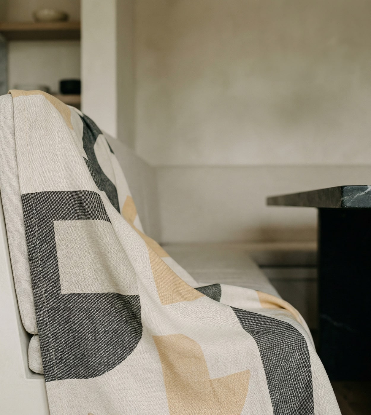 Gemini said A close-up, angled shot of a heavy woven fabric draped over the back of a light-colored banquette seat. The fabric features a bold, large-scale geometric pattern in charcoal grey, muted ochre, and off-white. In the background, a dark, textured stone table sits in a minimalist room with soft beige plaster walls. The lighting is natural and soft, highlighting the coarse texture of the textile and the clean lines of the interior design.