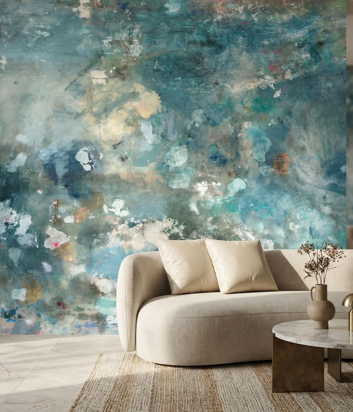 A contemporary living room featuring blue distressed abstract wallpaper with layered painterly textures. A curved beige sofa with cushions sits beside a marble coffee table with a ceramic vase and dried flowers, creating a calm and artistic interior.