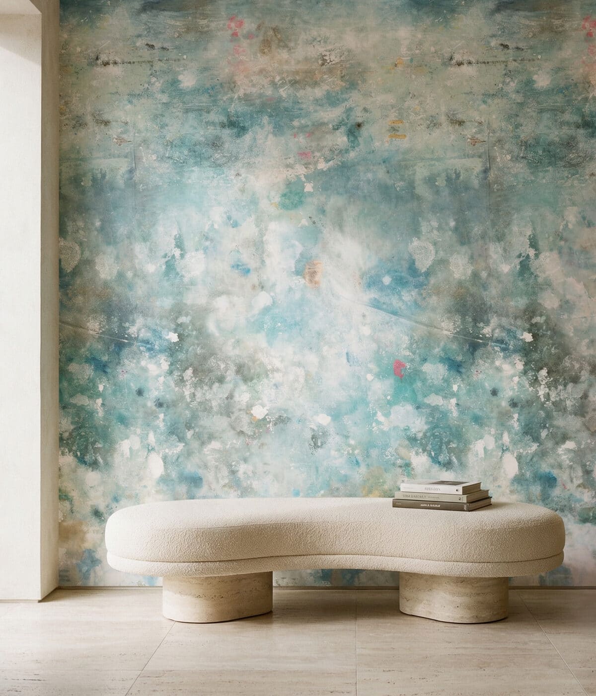 A modern room defined by a large-scale abstract wallpaper in shades of teal, blue, and white with a distressed, watercolor-like texture. Centered against the wall is a curved, cream-colored bouclé bench supported by two thick, cylindrical stone bases. A few books are neatly stacked on one end of the bench, and the space is finished with neutral-toned stone floor tiles and soft, natural lighting.