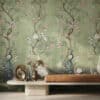 A stylish interior featuring a long stone bench topped with a caramel-brown leather cushion. Decorative objects—including a brass sculptural ornament and small metal vases with dried flowers—are arranged on the bench. The wall behind is covered in a green chinoiserie-style wallpaper depicting flowering tree branches, delicate blossoms, butterflies, and birds, including peacocks and a white crane, creating a lush, elegant garden scene.