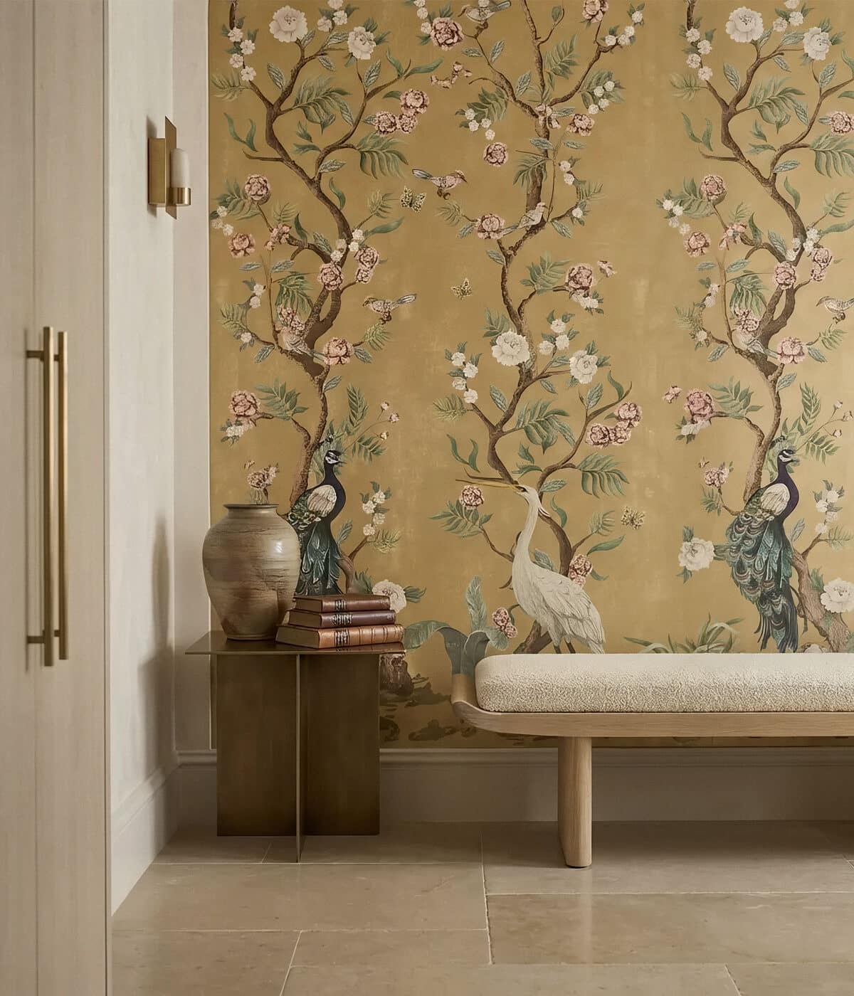 An elegant interior corner featuring a light wood bench with a cream upholstered cushion beside a small dark metal side table holding stacked books and a ceramic vase. The wall behind is covered in a gold chinoiserie-style wallpaper mural depicting flowering tree branches, delicate blossoms, butterflies, and birds including a peacock and a white crane. The warm gold background creates a rich, refined atmosphere.