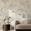 Elegant living room featuring a vintage chinoiserie-style wall mural with blue herons and delicate flowering branches on a warm beige background. In front of the mural sits a curved neutral sofa and a round side table with a brass bird sculpture and a small vase of dried flowers, creating a refined, serene atmosphere.
