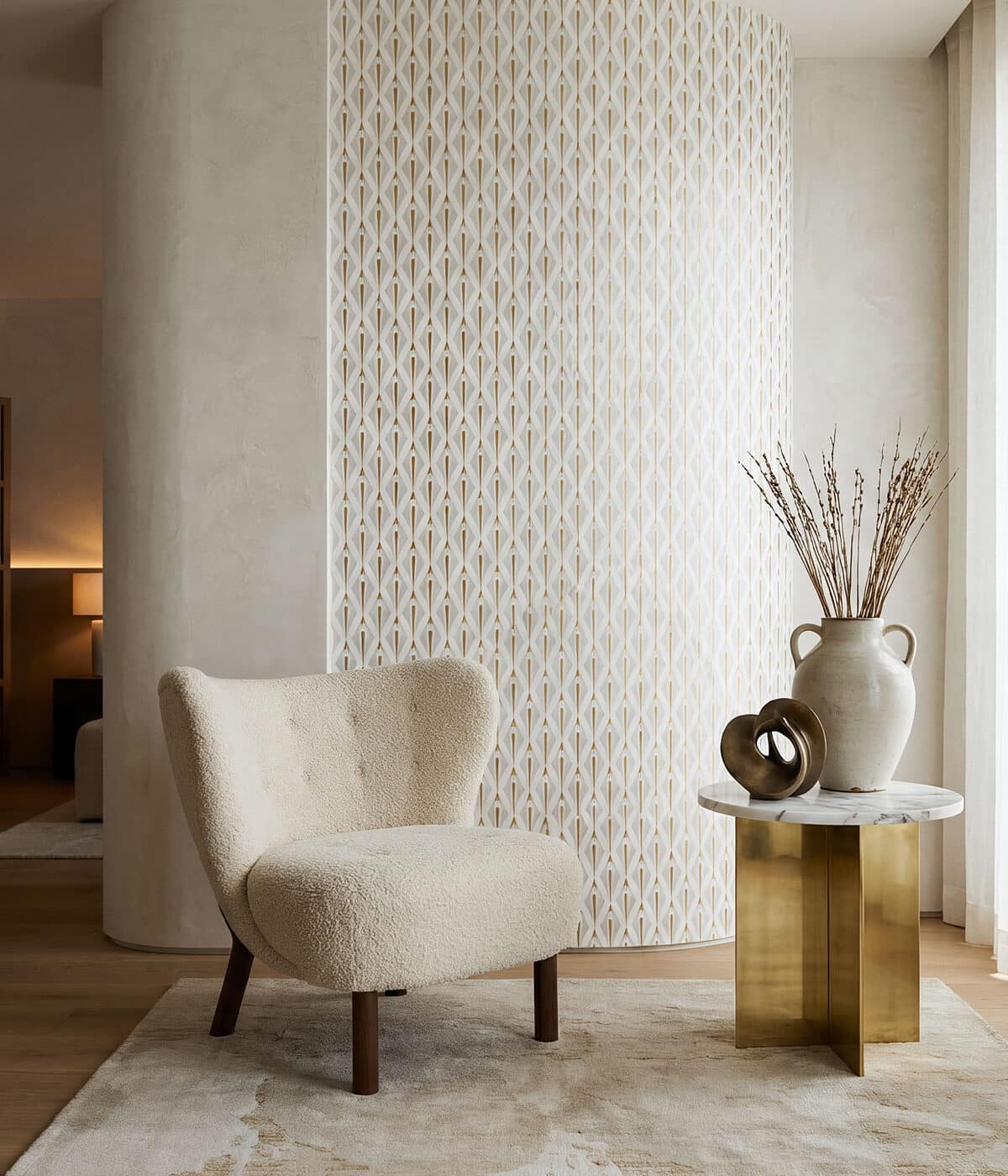 A refined, neutral-toned interior featuring a curved wall with a subtle geometric wallpaper pattern in soft white and beige. In the foreground sits a cream boucle accent chair with dark wooden legs, placed on a light textured rug. Beside it is a small round marble-top side table with a brushed brass base, styled with a ceramic vase holding dried branches and a sculptural decorative object. Sheer curtains filter natural light, creating a warm and elegant atmosphere.
